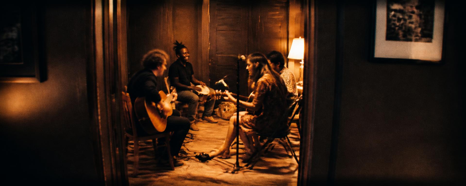 Músicos tocando juntos em uma sala, vistos de longe através de uma porta